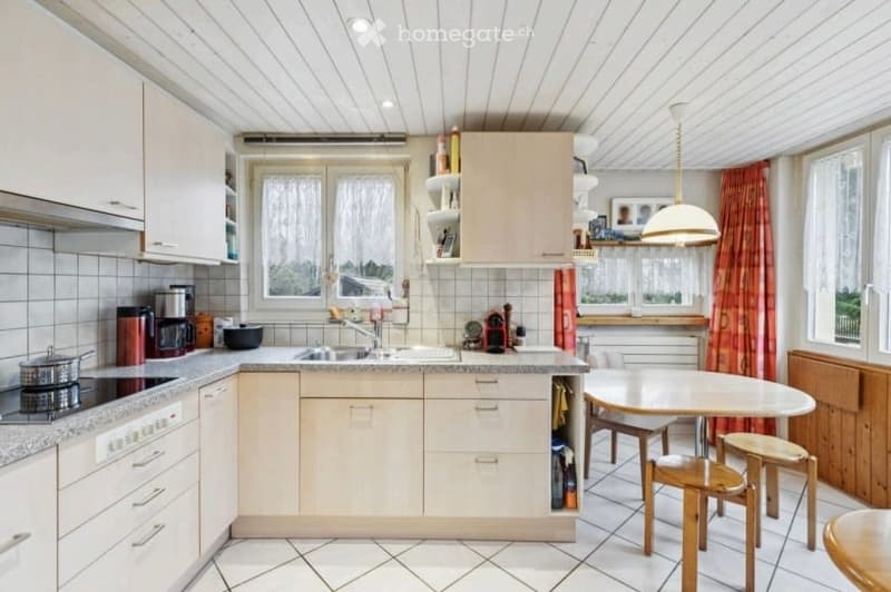 Bright kitchen with white cabinets, tiled walls, and a central island. Includes a sink, stove, coffee maker, and a small dining area with a table and stools.