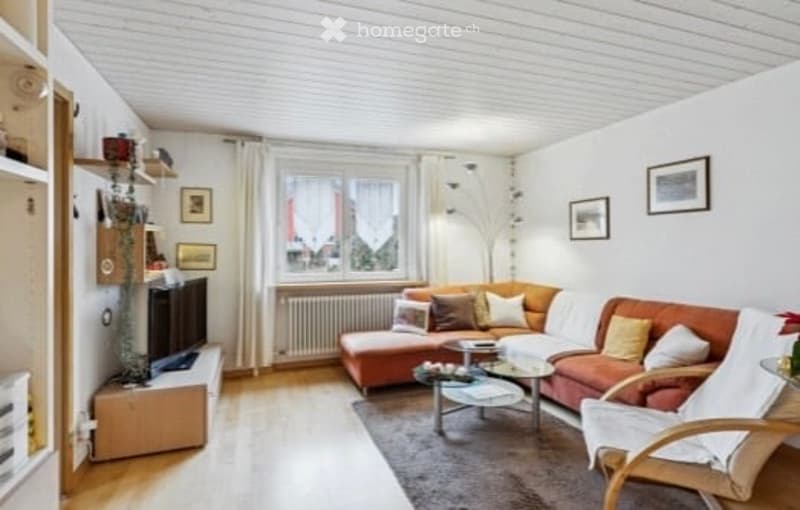 Modern living room with white walls, wooden flooring, large window, grey sofa, coffee table, armchair, TV stand, and decorative picture frames.