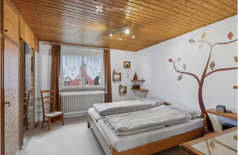 Cozy bedroom with wooden ceiling, two single beds, a wooden chair, nightstand, radiator, and decorative shelves.