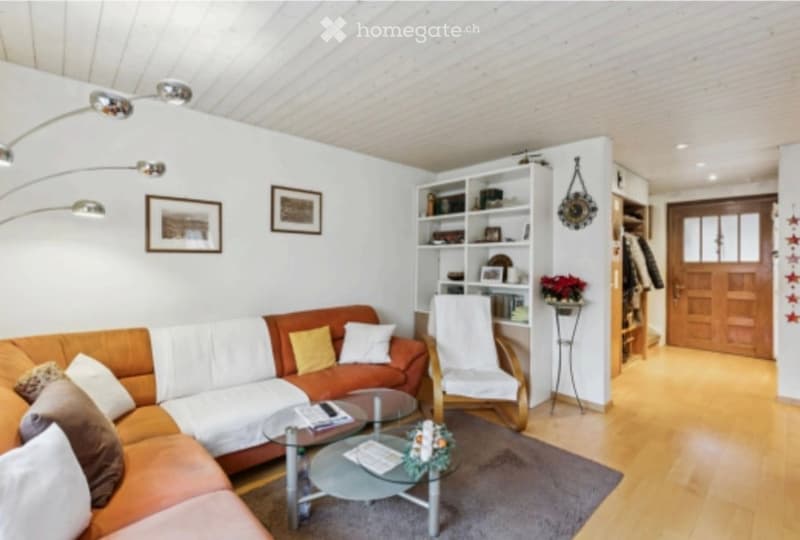 Living room with a wooden floor, white walls, and a modern design. Features include a sectional sofa, glass coffee table, white armchair, bookshelf, and decorative items.