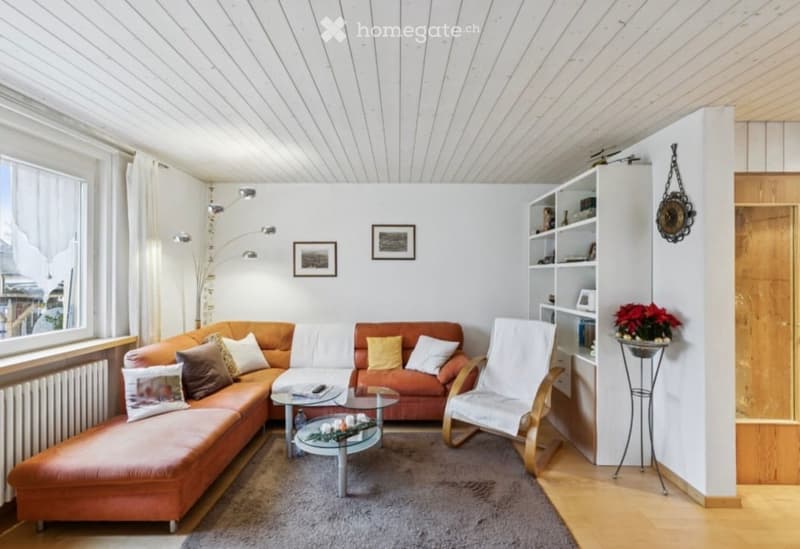 Modern living room with a large window offering a view, featuring a sectional sofa, armchair, glass coffee table, bookshelf, and decorative items.