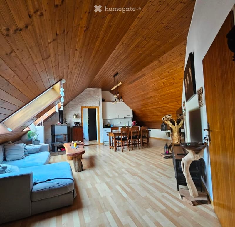 Open-plan living and dining area with wooden ceiling and floor, fireplace, sofa, coffee table, dining table with chairs, and kitchen area with cabinets.