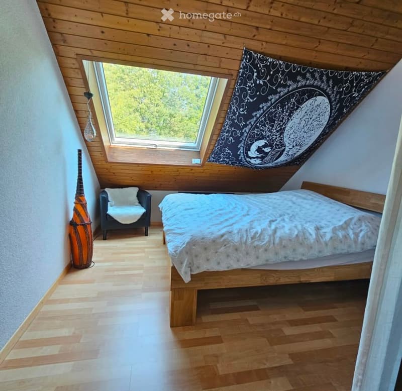 Cozy attic bedroom with wooden slanted ceilings, skylight windows, a bed, a small chair, a decorative vase, and a wall hanging.
