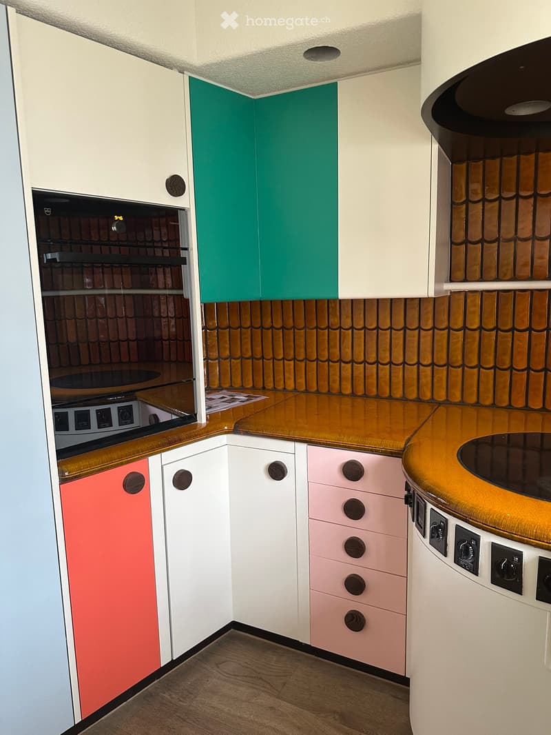 A compact kitchen with colorful cabinets in white, pink, and green, wooden countertops, and built-in appliances including an oven and stove.