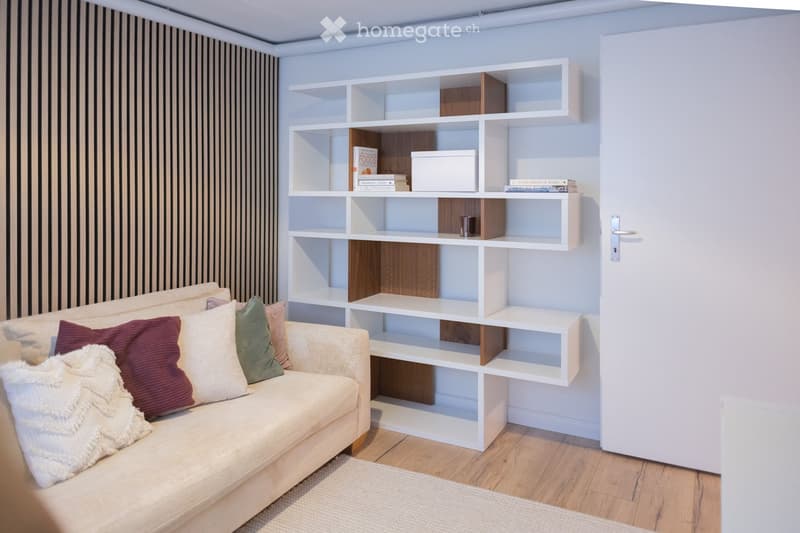 A modern living room with a light-colored sofa adorned with multiple pillows, a white modular bookshelf filled with books and a cup, and a wooden floor.