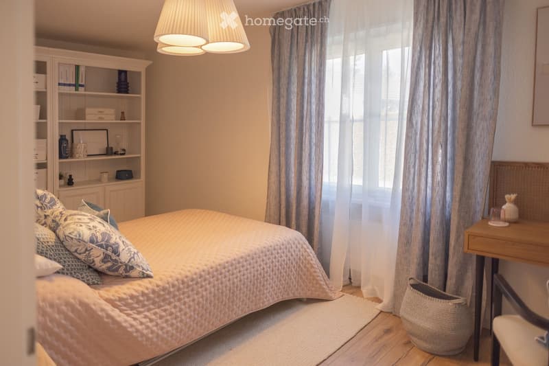 Cozy bedroom with a large bed, patterned pillows, a wooden nightstand, a woven basket, a shelf with decorative items, and a large window with curtains.