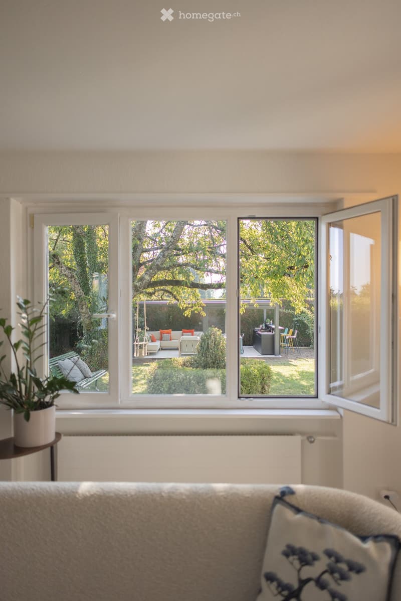 Living room with large windows overlooking a garden, featuring a sofa, a potted plant on a side table, and a bench visible through the window.