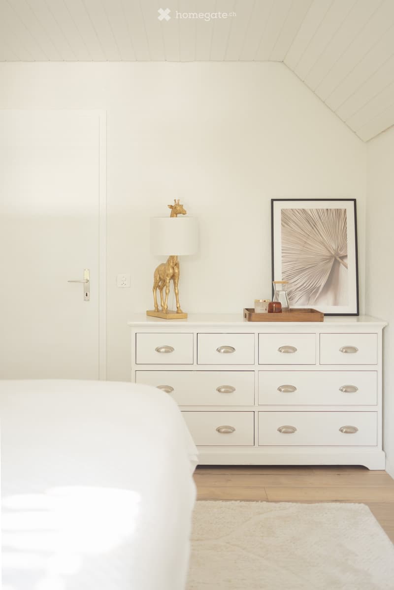 A modern bedroom with a white nightstand, a lamp, a framed picture, a decorative tray, and a giraffe figurine. The room features a white bed and a light-colored rug on a wooden floor.