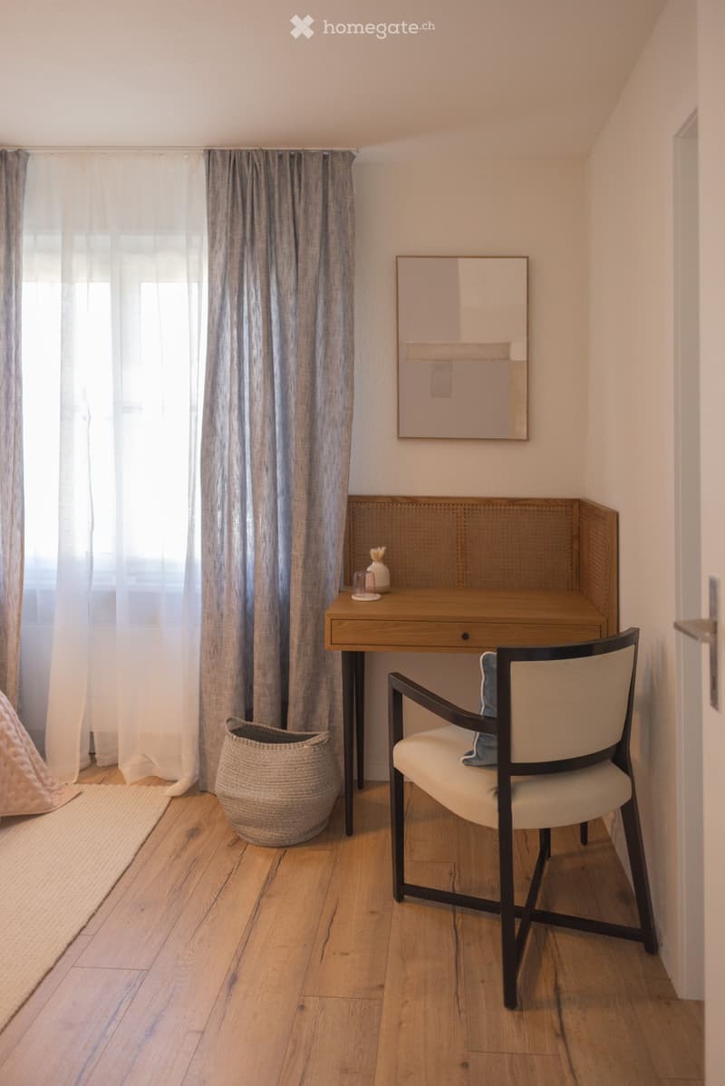 A cozy, well-lit room with wooden flooring, a large window with curtains, a desk with a chair, a woven basket, and a mirror on the wall.