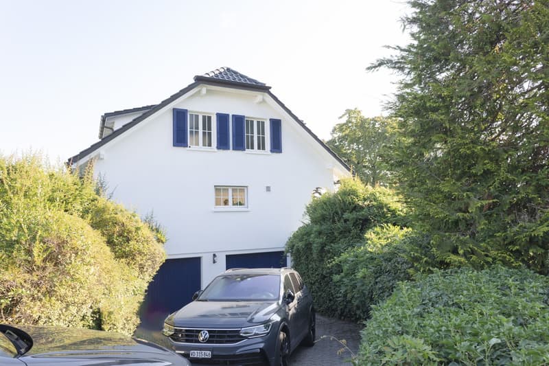 Two-story white house with blue shutters and garage, surrounded by greenery and parked car.