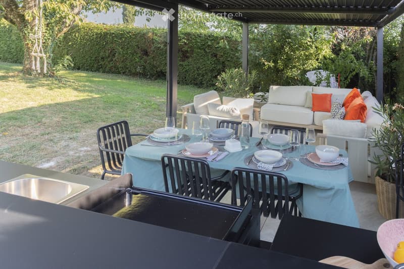 A covered outdoor patio with a dining table set for six, surrounded by a sofa and chairs, with a lush green garden in the background.