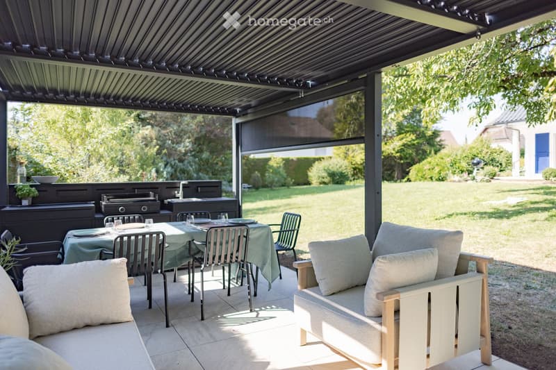 A covered outdoor patio with a sofa, dining table, chairs, and a barbecue area, surrounded by greenery and a lawn.