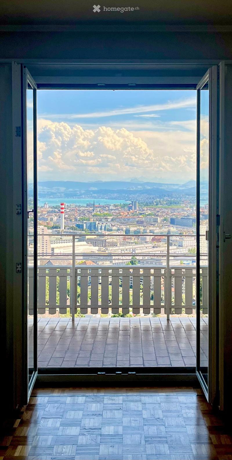Balcony with wooden floor and railing, offering a panoramic view of a cityscape, mountains, and a body of water.