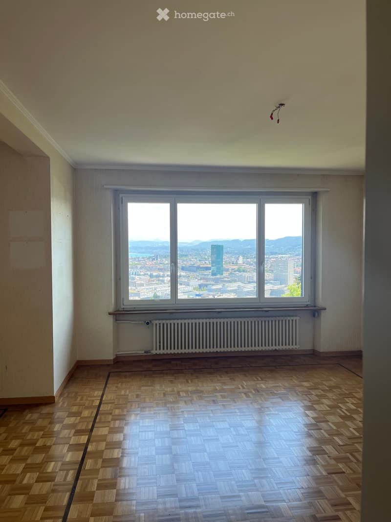 Empty room with wooden flooring, large window offering a cityscape view, radiator below the window, and neutral-colored walls.