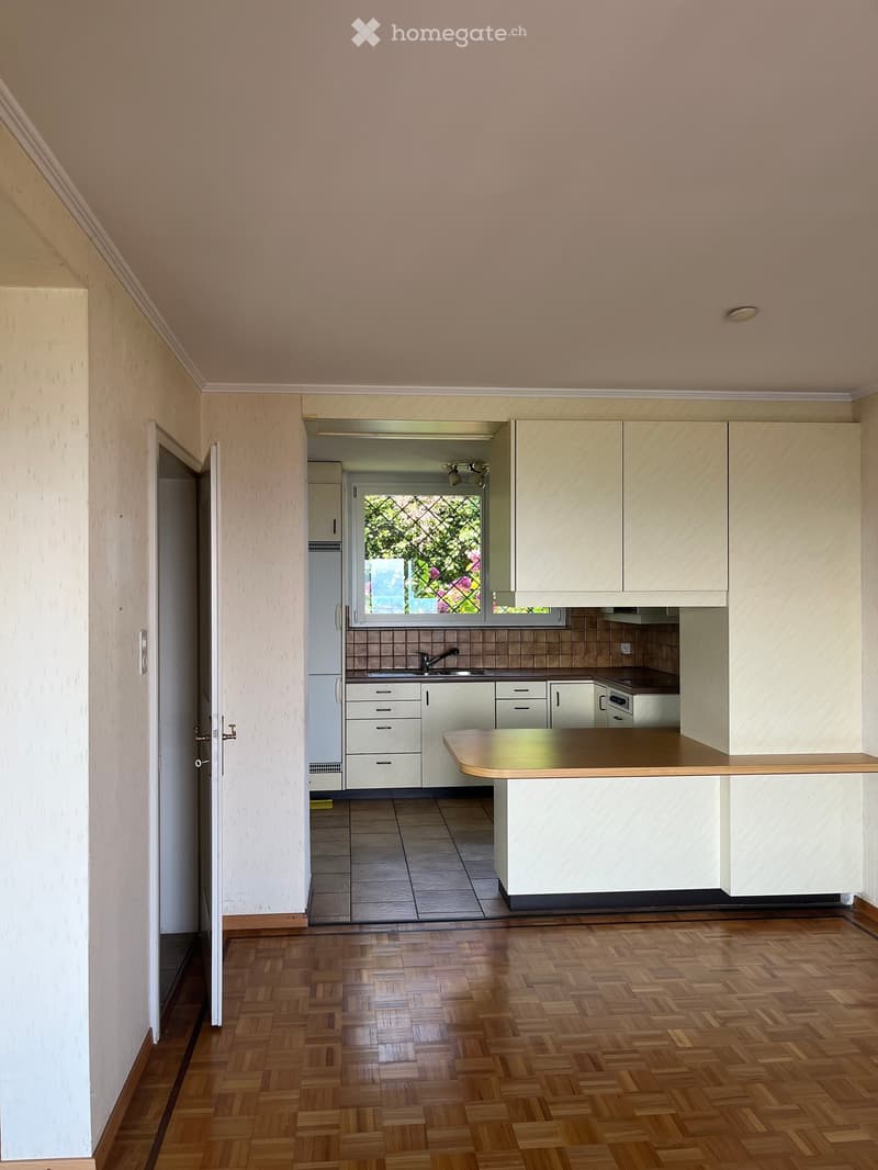 Bright kitchen with light-colored cabinets, a central island, and a window overlooking a garden. The floor is wooden, and the walls are painted in a soft beige tone.