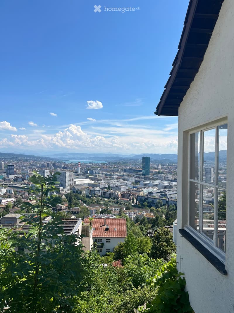 Panoramic city view from a balcony with clear blue skies, distant mountains, and urban skyline.