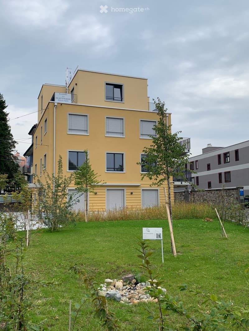 A modern yellow building with multiple windows and balconies, surrounded by a green lawn and small garden area.