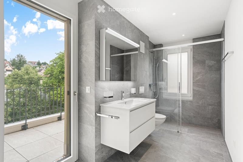 Modern bathroom with grey tiled walls, white sink, mirror, and a view of greenery through the open balcony door.