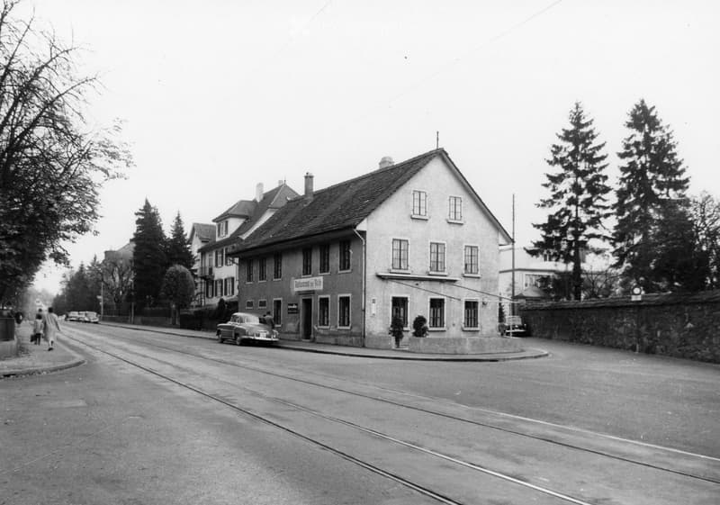 A two-story building with a pitched roof and multiple windows, located on a street with a few parked cars and pedestrians nearby.