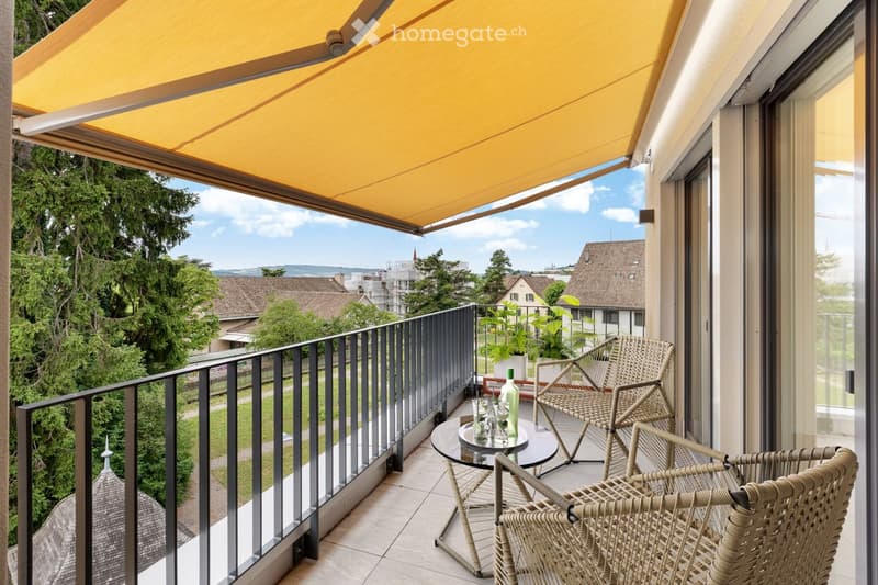 Balcony with a yellow retractable awning, glass railing, and outdoor furniture including a round table, chairs, and a bottle on the table. The view shows a green landscape with trees and buildings.