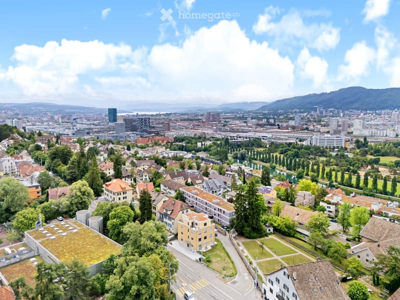 Aerial view of a suburban neighborhood with residential buildings, trees, and a road network, with a cityscape and mountains in the background.