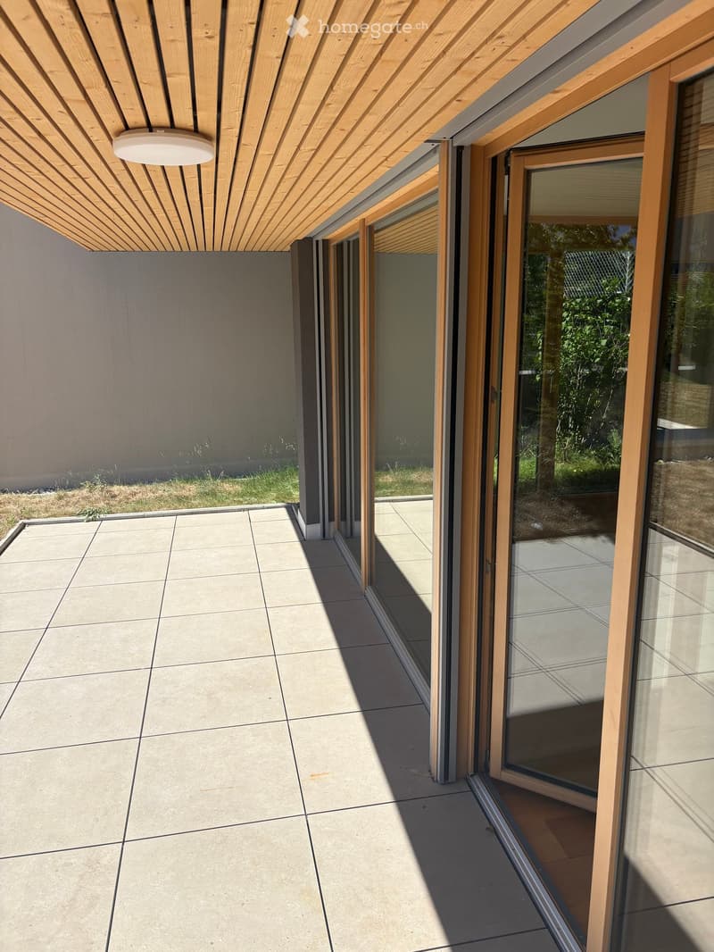 Modern balcony with large glass sliding doors, wooden ceiling, tiled floor, and view of greenery outside.