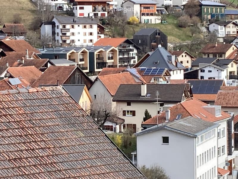 A panoramic view of a residential area with multiple houses featuring varied roof styles, including red-tiled and grey roofs, some with solar panels, nestled in a hilly landscape.