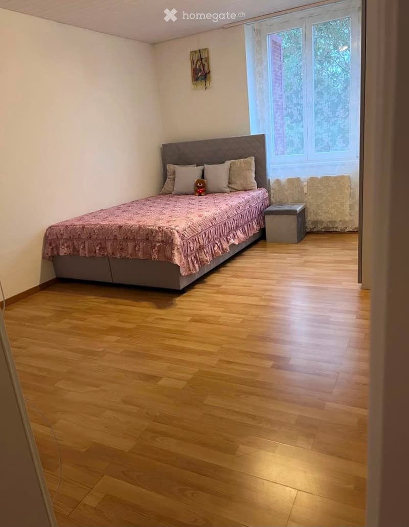 Cozy bedroom with wooden flooring, a neatly made bed with pillows and a decorative item, a small stool, and a framed picture on the wall.