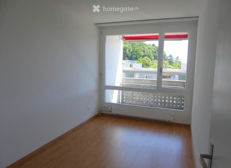 Empty room with wooden flooring, white walls, and large windows offering a view of greenery outside.