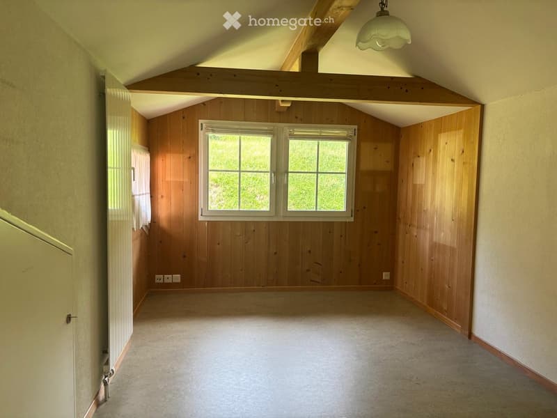 Empty attic space with wooden walls, exposed wooden beams, and a large window letting in natural light. The floor is concrete, and there are electrical outlets visible on the walls.