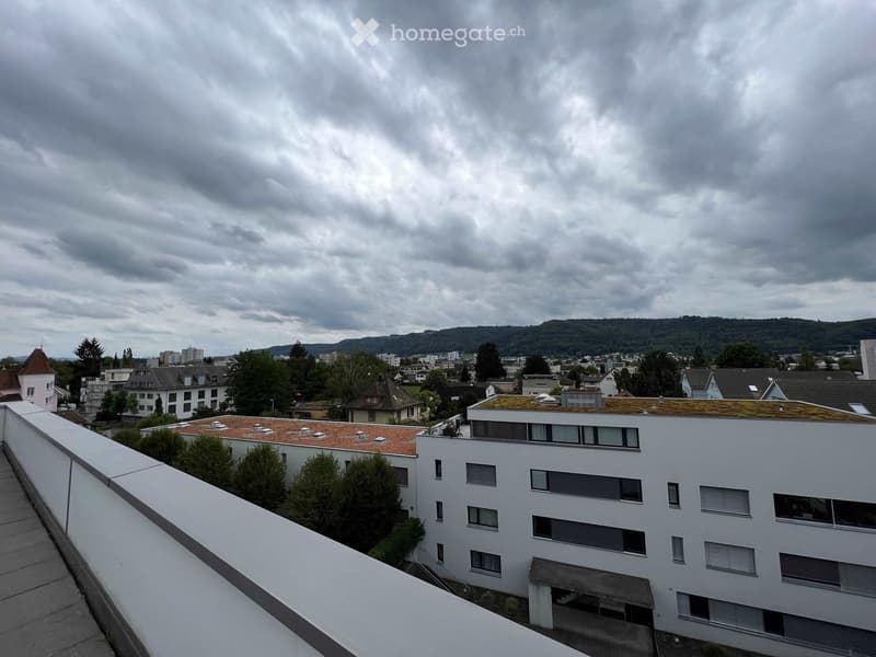 Balcony overlooking a residential area with multiple buildings, trees, and distant hills under a cloudy sky.