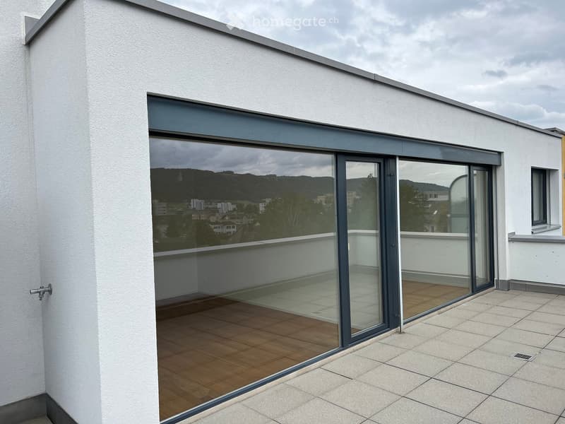 Modern balcony with large glass sliding doors, wooden decking, and tiled floor, overlooking a cityscape with hills in the background.