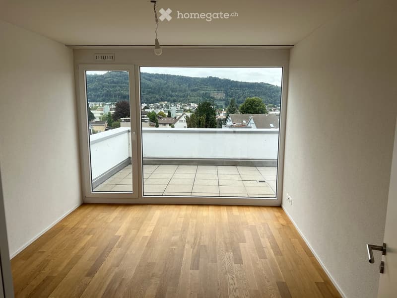 Empty room with wooden flooring, large sliding glass doors leading to a balcony, and a scenic view of a town and hills.