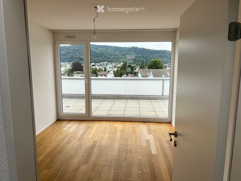 Empty room with wooden flooring, large windows, and a view of a cityscape and greenery. The room has a sliding glass door leading to a balcony.