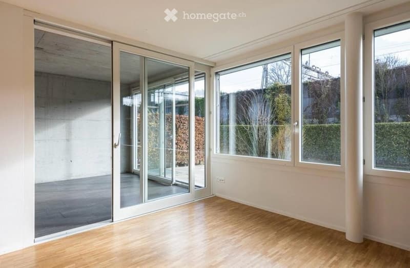 Empty room with wooden flooring, large windows, and sliding glass doors leading to an outdoor area.