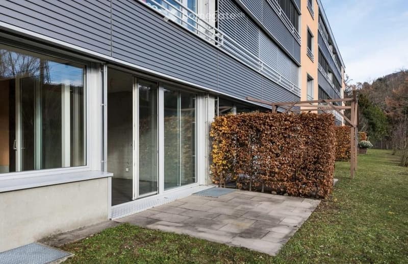 Modern multi-story building with large glass windows and sliding doors, surrounded by neatly trimmed hedges and a paved pathway.