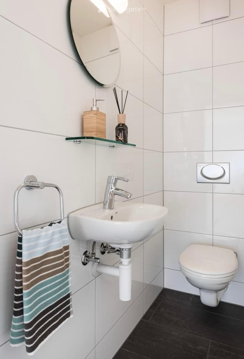 Modern bathroom with a white sink, chrome faucet, toilet, towel on a rack, and a glass shelf holding a soap dispenser and decorative items.