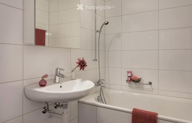 A modern bathroom with white tiled walls and floor, featuring a sink with a chrome faucet, a bathtub with a handheld shower, a mirror, and decorative items including a flower vase and a towel.