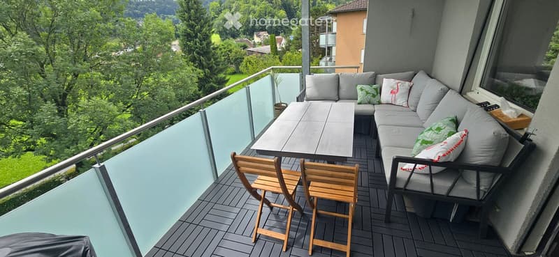 Balcony with a grey sectional sofa, wooden table, and two wooden chairs, overlooking a lush green landscape.