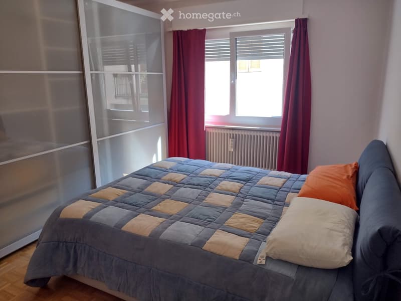 A modern bedroom with a large bed covered in a blue and beige quilt, a sofa with pillows, and large windows with red curtains.