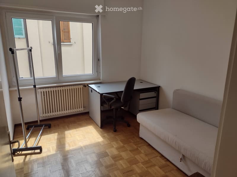 A small, well-lit room with wooden flooring, a sofa, a desk with a chair, and a radiator under the windows.