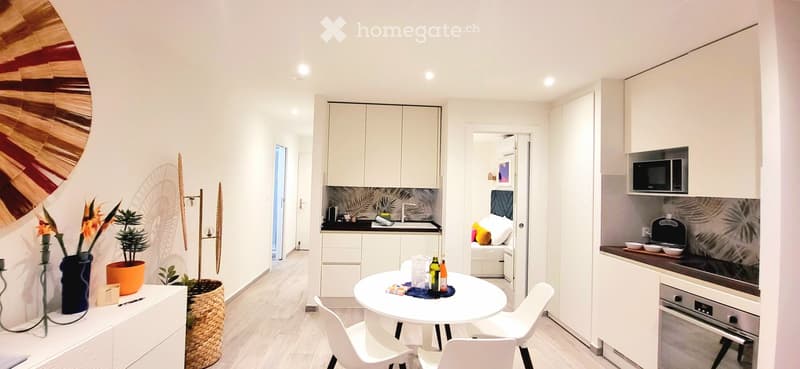 Modern, compact studio apartment with a combined living, dining, and kitchen area. Features include a white dining table with chairs, a small kitchen with cabinets, a sink, and appliances, and a cozy sleeping area with a bed and pillows.