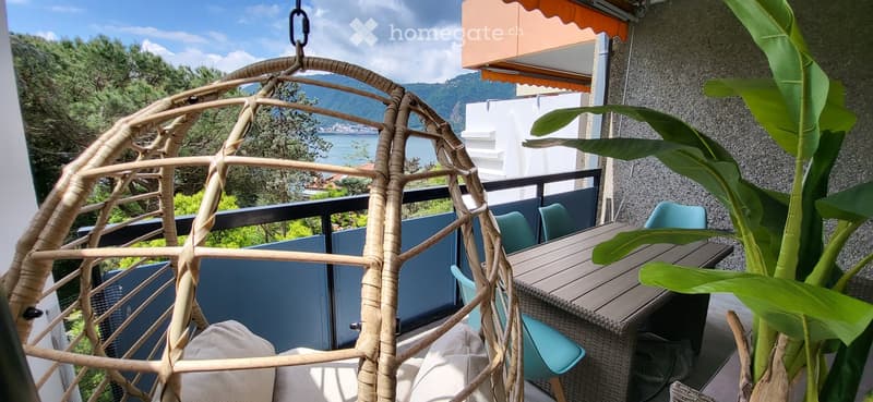 Balcony with a wicker hanging chair, a wooden table, turquoise chairs, and a large leafy plant, overlooking a scenic lake and mountains.