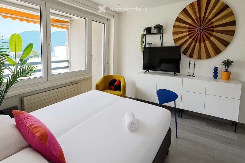 Bright bedroom with a white bed, colorful pillows, a yellow armchair, a white TV cabinet, and a large window offering a scenic view.