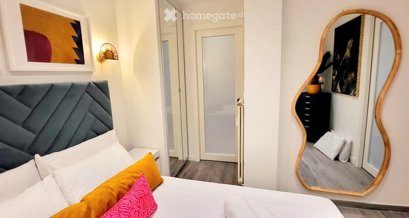 Modern bedroom with a white bed, grey headboard, colorful pillows, a large wooden-framed mirror, and a nightstand with a lamp.