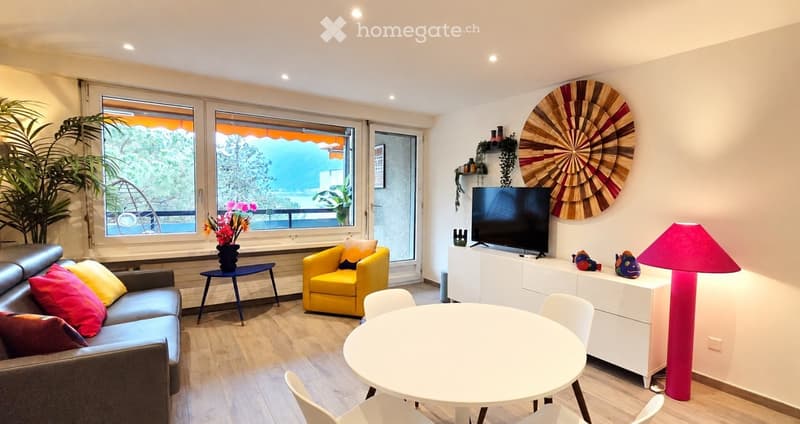 Bright living room with large windows offering a view, featuring a grey sofa, yellow armchair, white dining table, floor lamp, TV, and decorative wall piece.