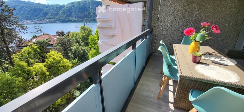 Balcony with a scenic view of a lake and mountains, featuring a small table with chairs, a vase with pink flowers, and a small potted plant.