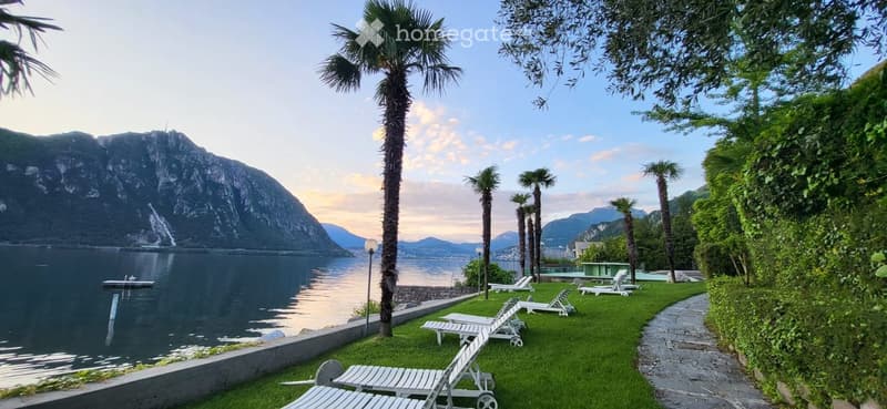 Lush garden area with lounge chairs, palm trees, and a scenic lake view with mountains in the background.
