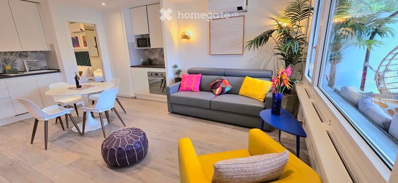 Modern living room with a grey sofa, colorful cushions, a yellow armchair, a round white dining table with chairs, a blue side table, and a view of the ocean through large windows.