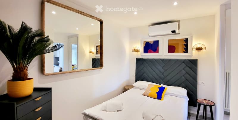 Modern bedroom with a grey upholstered headboard, white bedding, decorative pillows, a large mirror, a potted plant, and wall-mounted wall art.
